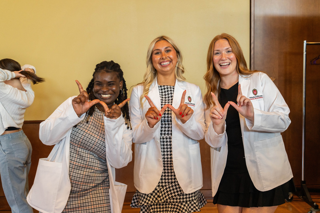 Three female students in white coats hold up "W" hands