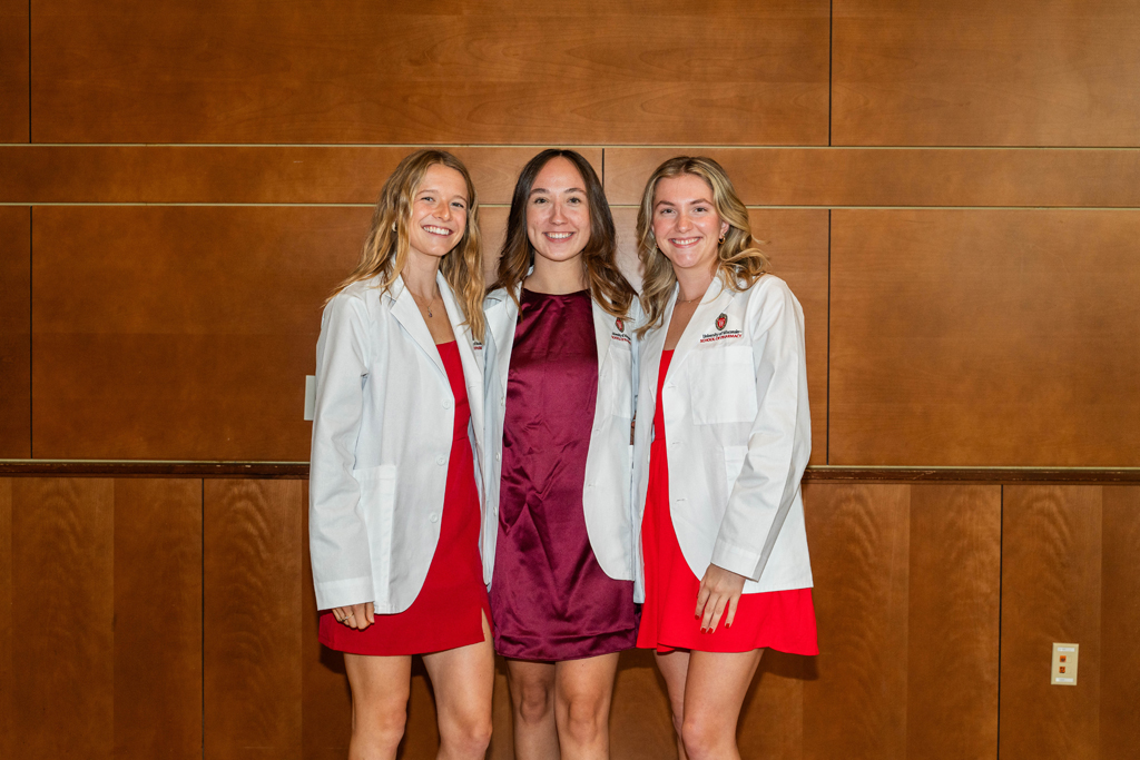 Three female students in white coats pose for a photo against a wooden wall