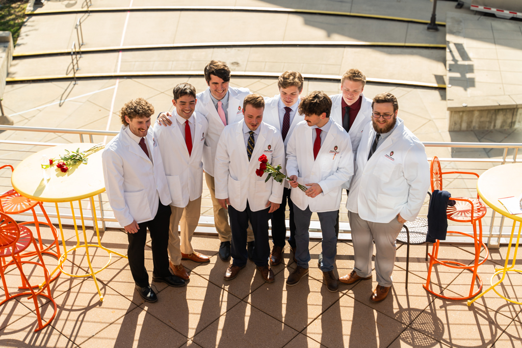 A group of male students in white coats gather for a photo. The photo is taken from above and the students are looking ahead of them toward a different, unseen photographer.