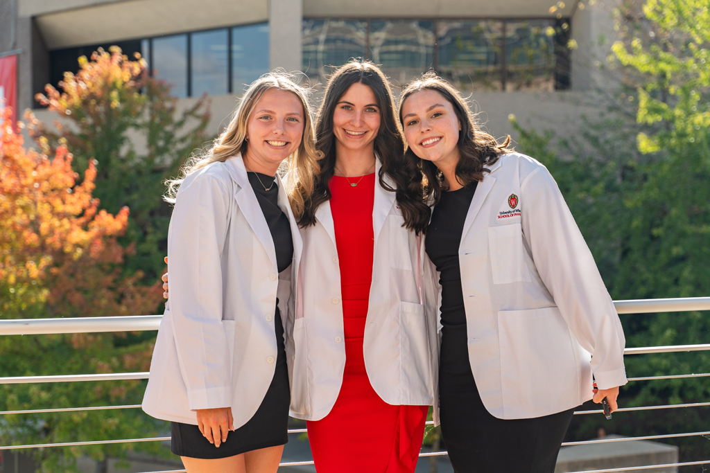 Three female students in white coats smile for a photo outside