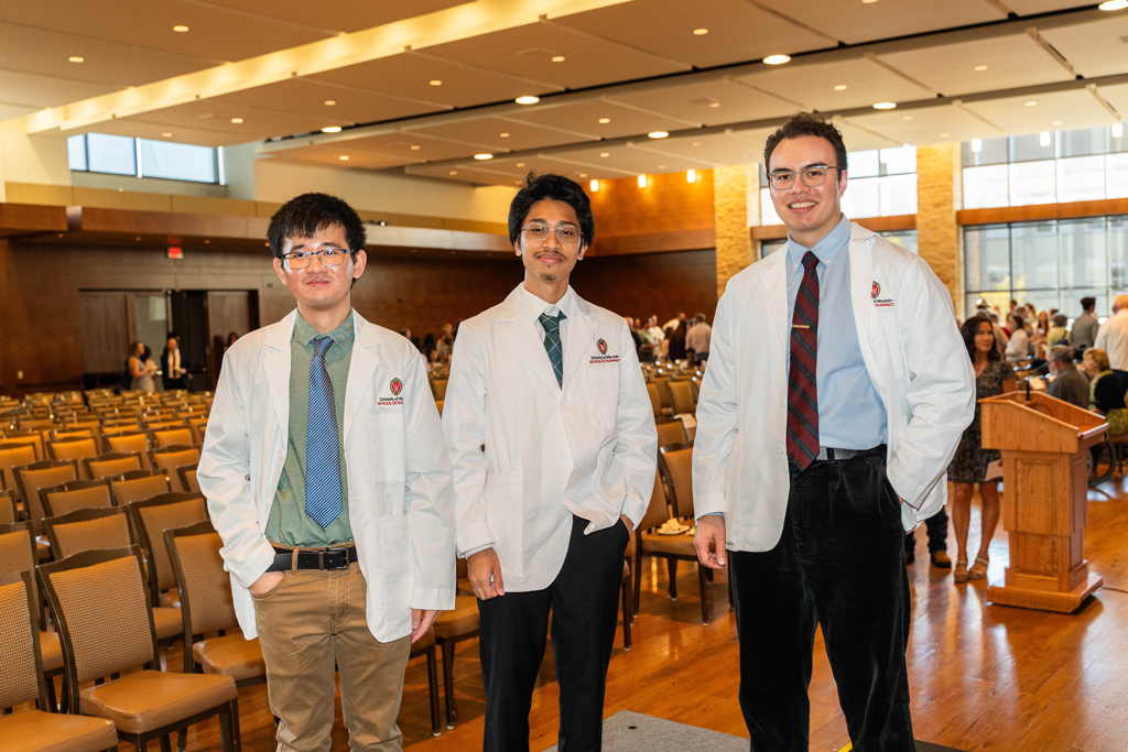 Three male students in white coats pose in front of empty chairs