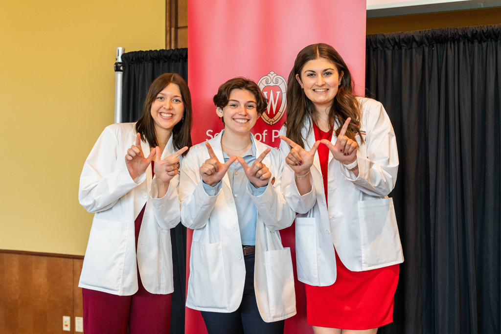 Three students in white coats pose in front of red School of Pharmacy banner