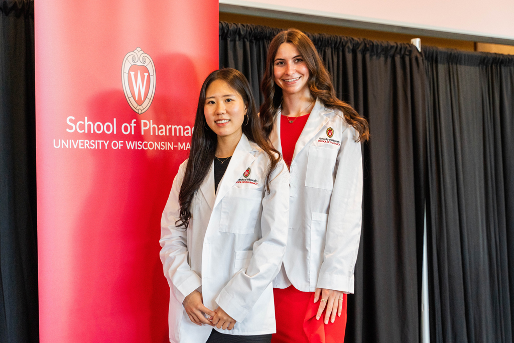 Two female students in white coats pose in front of a red School of Pharmacy banner