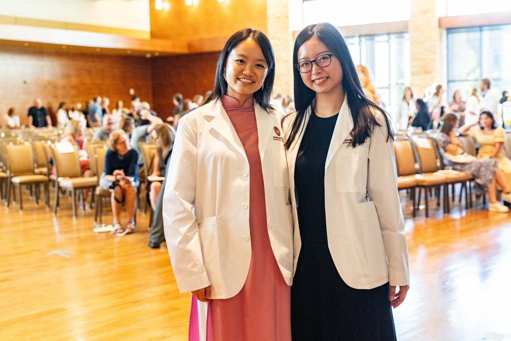 Two female students in white coats pose in front of audience chairs