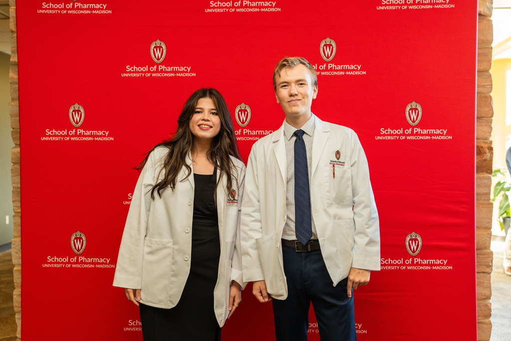 A female and male student in white coats pose in front of red School of Pharmacy background