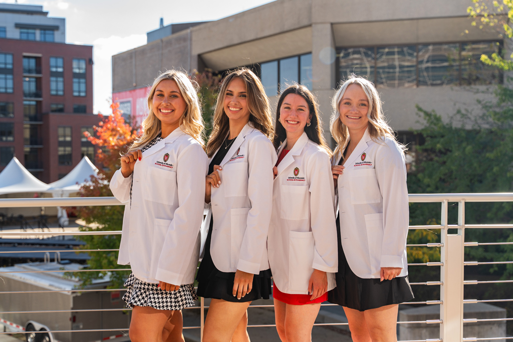 Four female students pose outside and hold their white coat lapel to showcase the School of Pharmacy logo