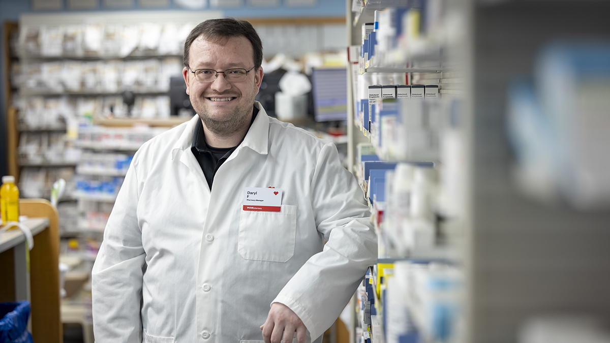 Daryl Fahrner poses in his white coat next to a shelf of medication