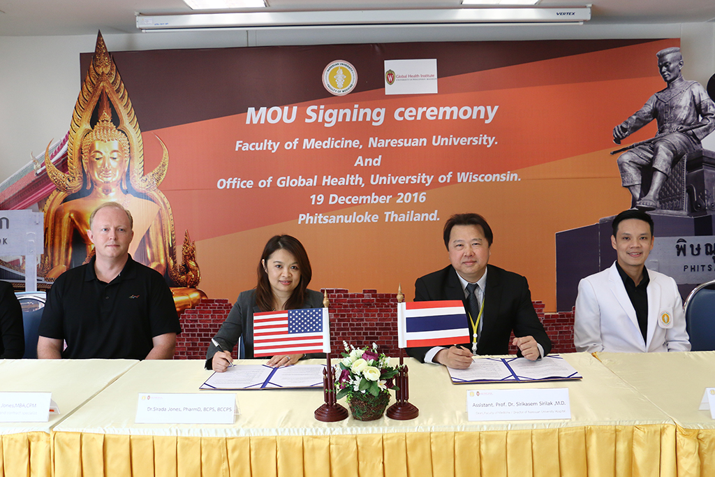 Four individuals seated at a table during an MOU signing ceremony between the Faculty of Medicine at Naresuan University and the Office of Global Health at the University of Wisconsin, held on December 19, 2016, in Phitsanuloke, Thailand. An American flag and a Thai flag are displayed on the table alongside flowers and signed documents.