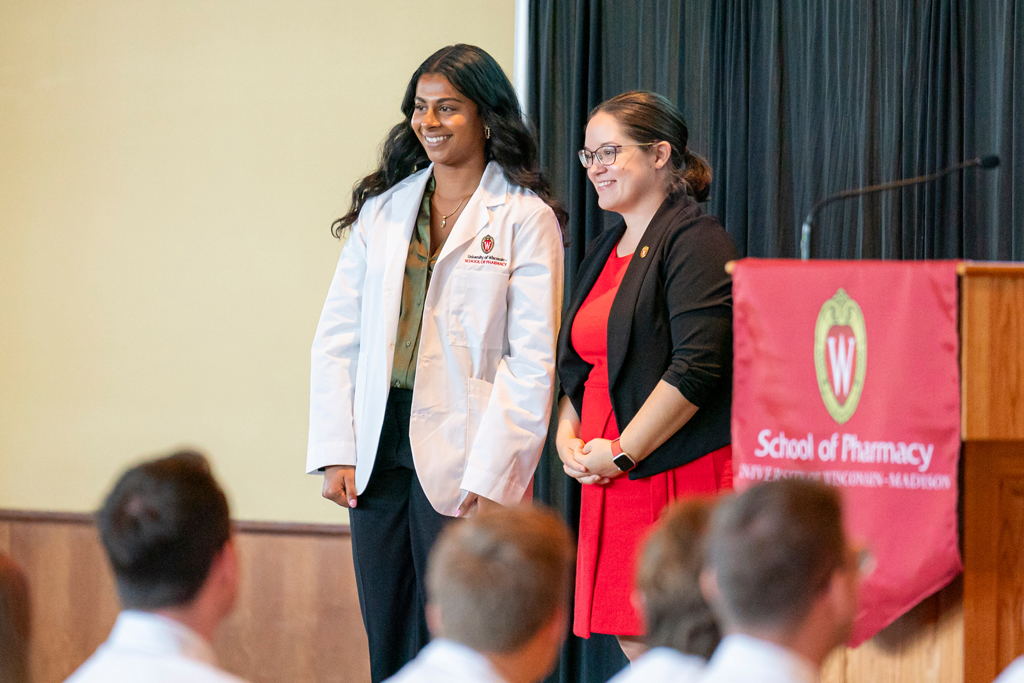 Student in white coat smiling with staff on-stage