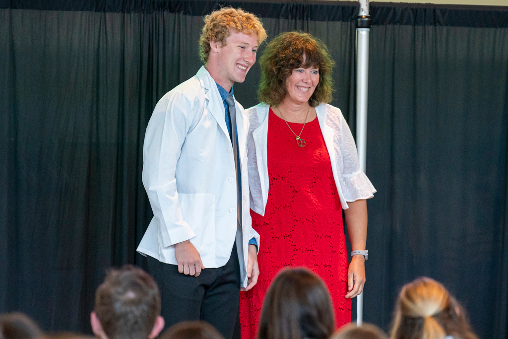 Student in white coat smiling with staff on-stage