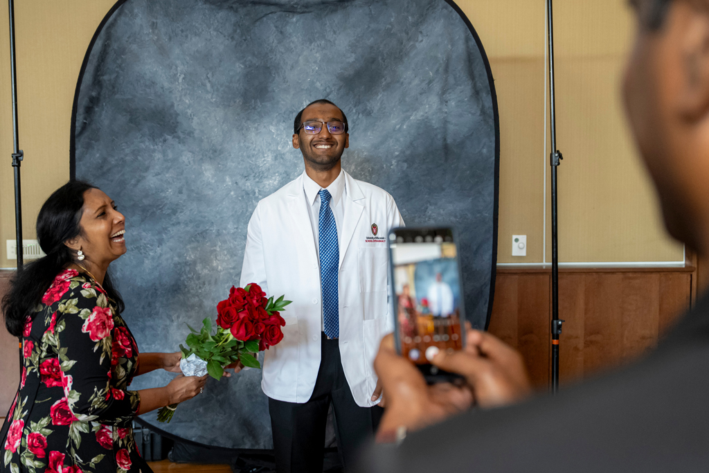 Student posing in white coat with his mom laughing from the side holding roses