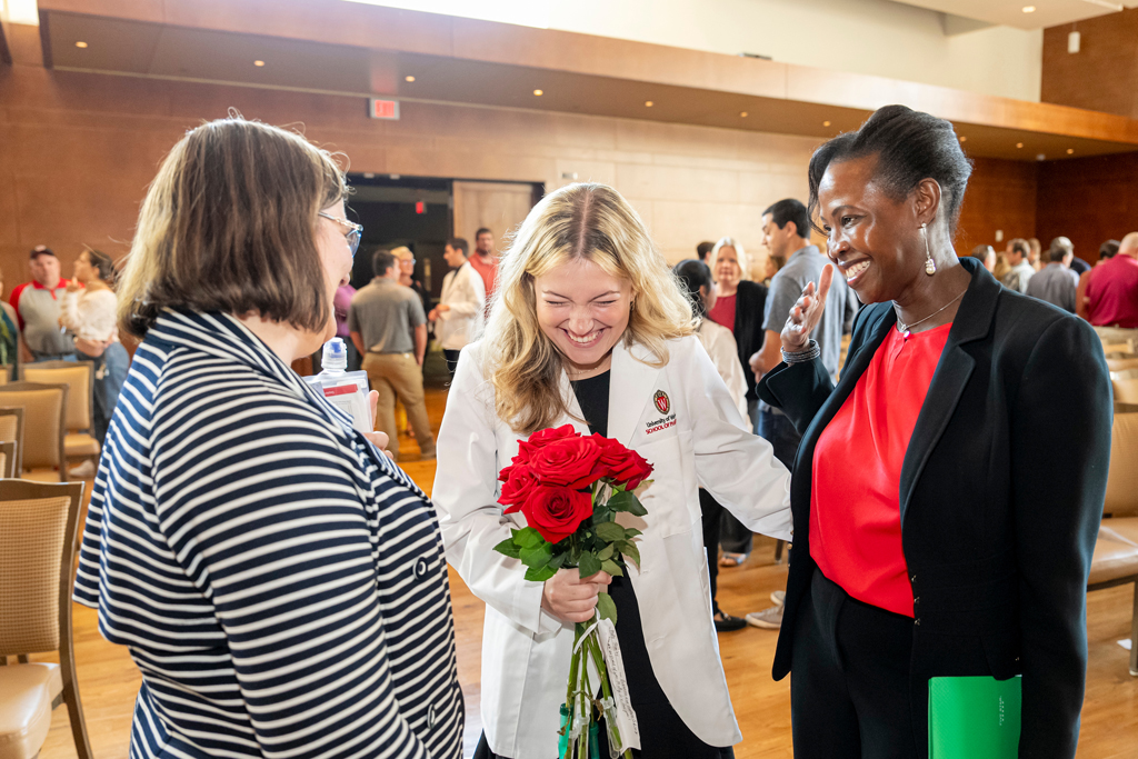 Student in white coat holding roses while laughing with her mother and a staff member