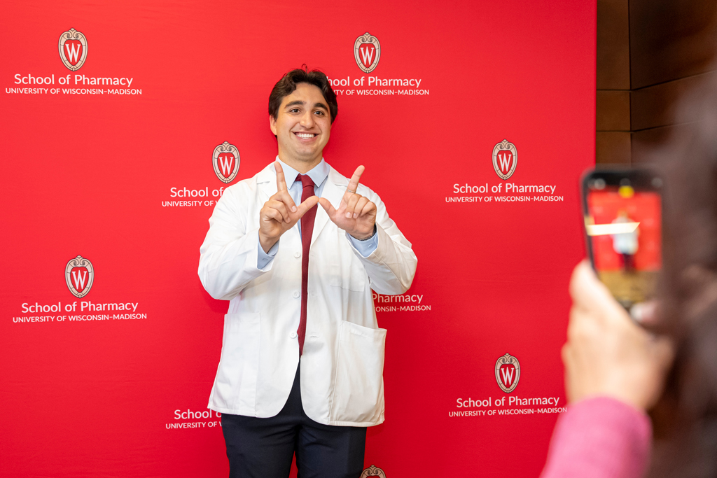 Student in white coat flashing "W" in front of School of Pharmacy background