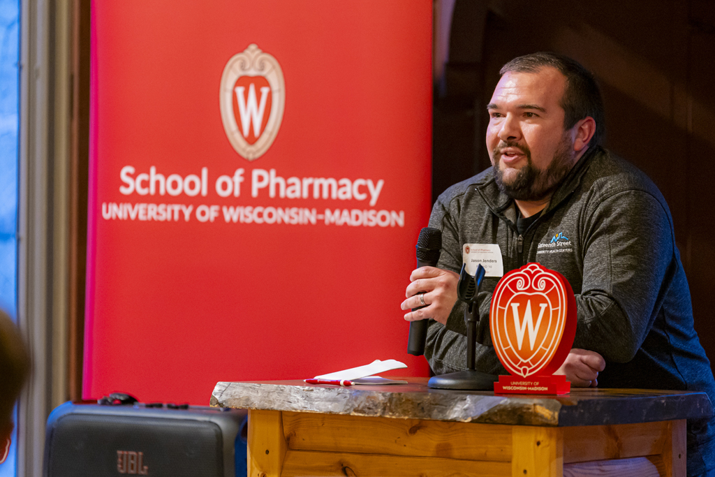 Jason Jenders speaks at a microphone in front of a School of Pharmacy banner