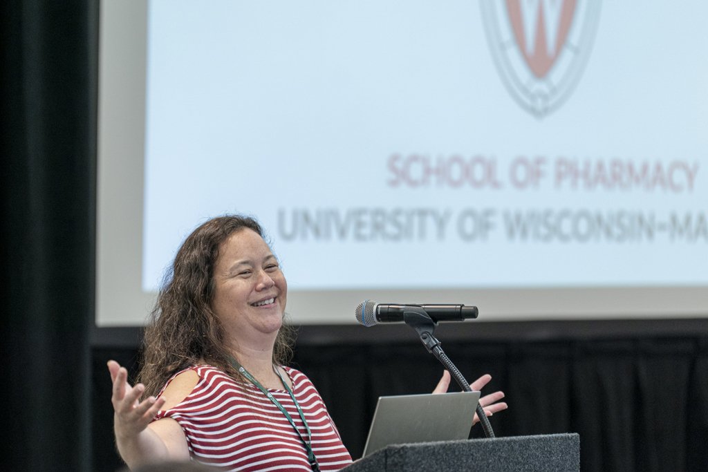 A woman in a striped shirt speaking at a microphone and gesturing with her hands