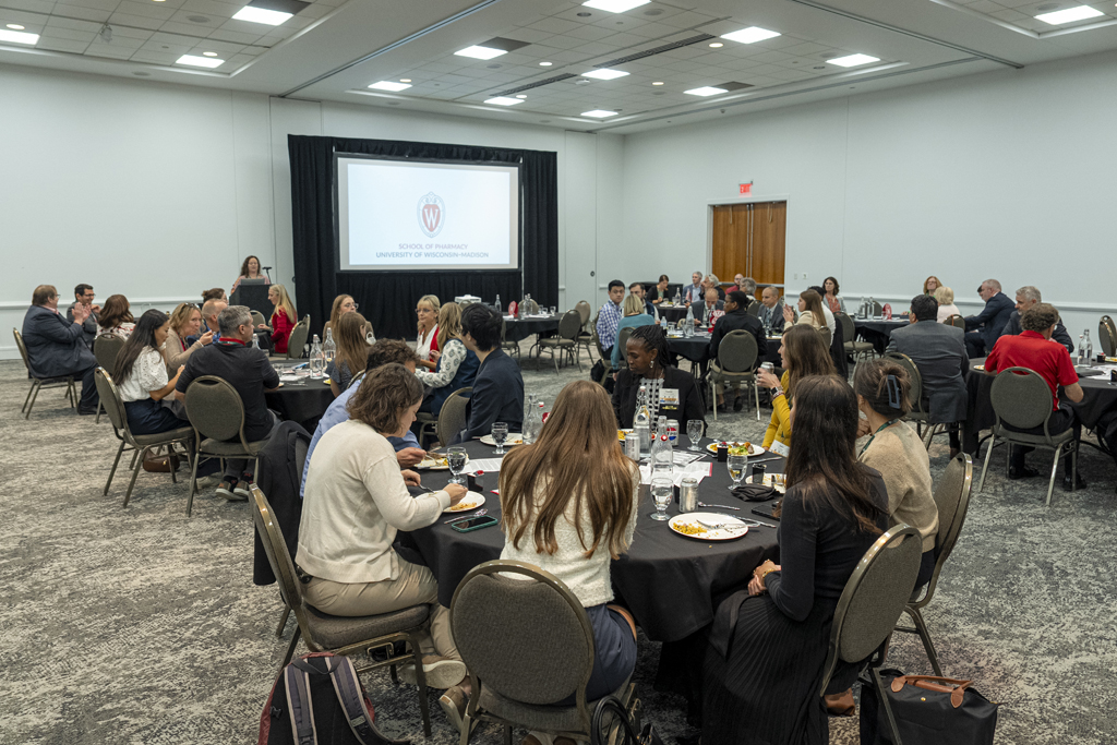 Wide shot of a large, well-lit conference room where people are seated at round tables covered with black tablecloths, eating dinner. On stage, a large screen displays the logo and text "SCHOOL OF PHARMACY, UNIVERSITY OF WISCONSIN-MADISON." A woman is speaking into a microphone on the left side of the stage.