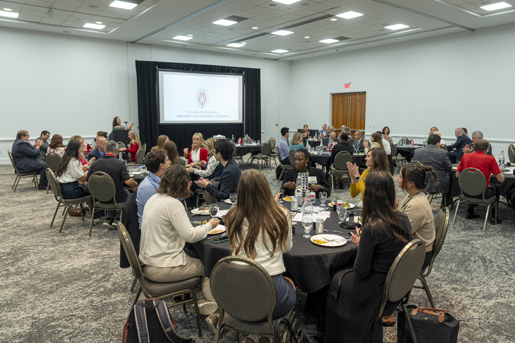 A wide shot of a large conference room with attendees seated at round tables, actively engaged in conversations while eating. The University of Wisconsin-Madison School of Pharmacy logo and name are projected onto a screen at the front of the room.