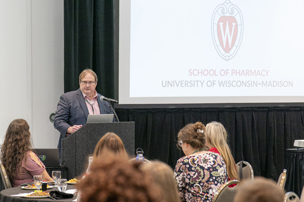 A man in a blue blazer and pink collared shirt is speaking from a podium to an audience seated at tables in a conference room. The University of Wisconsin-Madison School of Pharmacy logo and name are visible on the screen behind him.