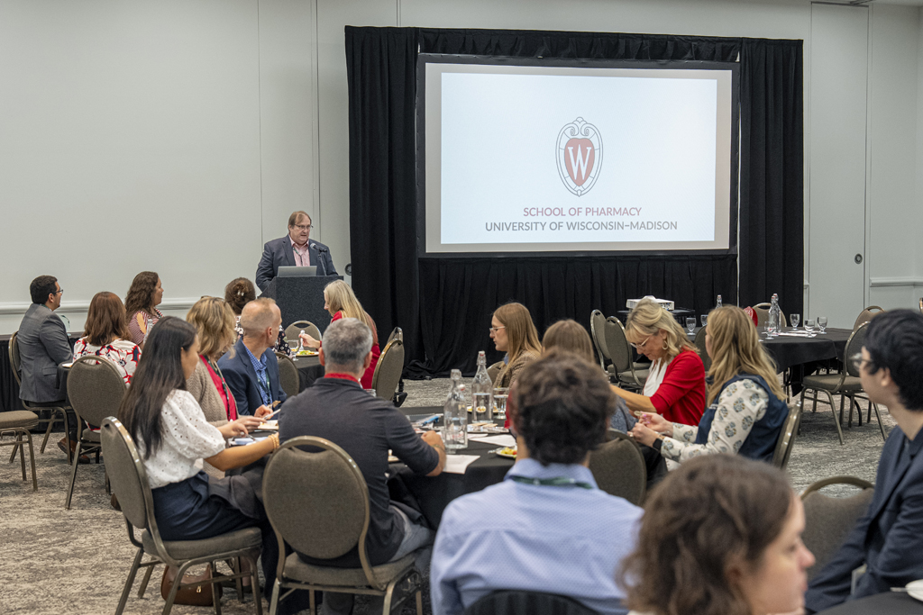 A man in a blue blazer is speaking from a podium at a formal gathering. The audience is seated at round tables, and the University of Wisconsin-Madison School of Pharmacy logo is projected onto the large screen.