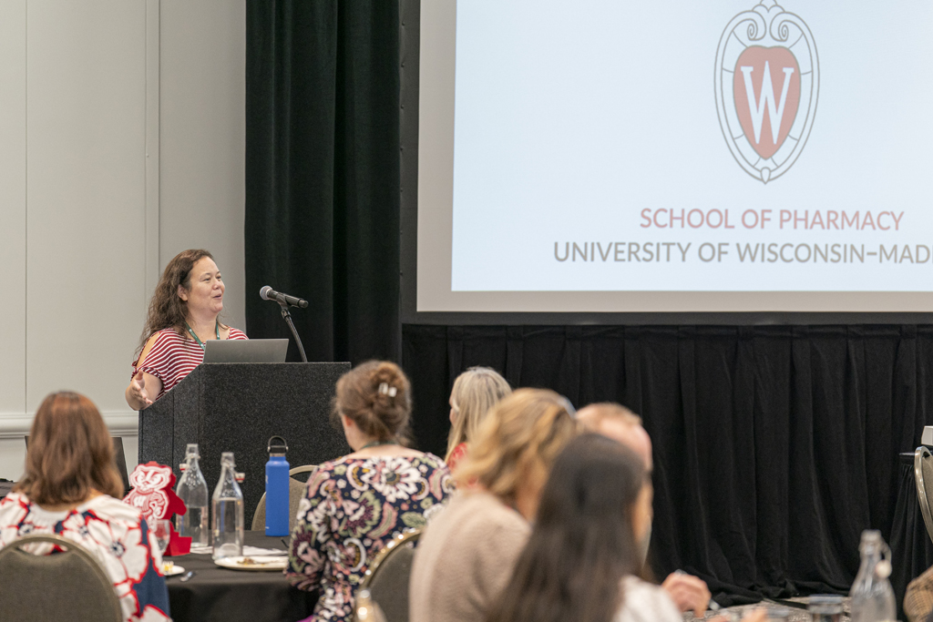A woman with long, curly hair and a striped shirt is smiling while speaking at a podium with a microphone. The University of Wisconsin-Madison School of Pharmacy logo and name are displayed on the screen behind her.