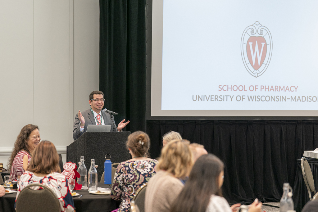 A man in a suit is speaking from a podium, gesturing with both hands. The University of Wisconsin-Madison School of Pharmacy logo and name are visible on the screen behind him.