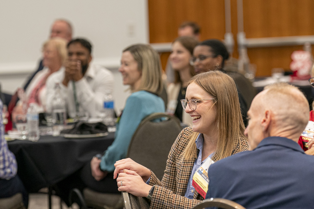 People in business casual attire site at round tables and listen to a speaker with smiles on their faces