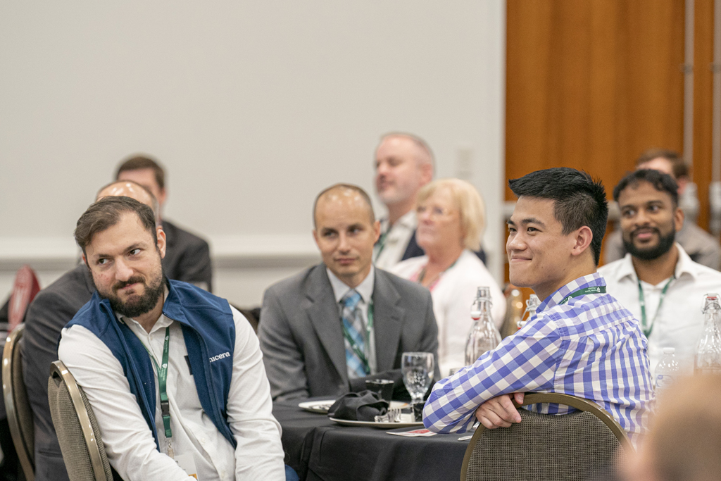 People in business casual attire site at round tables and listen to a speaker with smiles on their faces