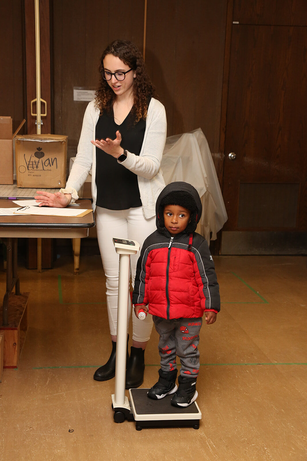 A young child in a winter coat stands on a scale near PharmD student Kathryn Frietag