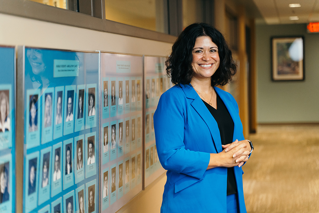 Maria Wopat stands in a hallway in front of black and white portraits on a blue background