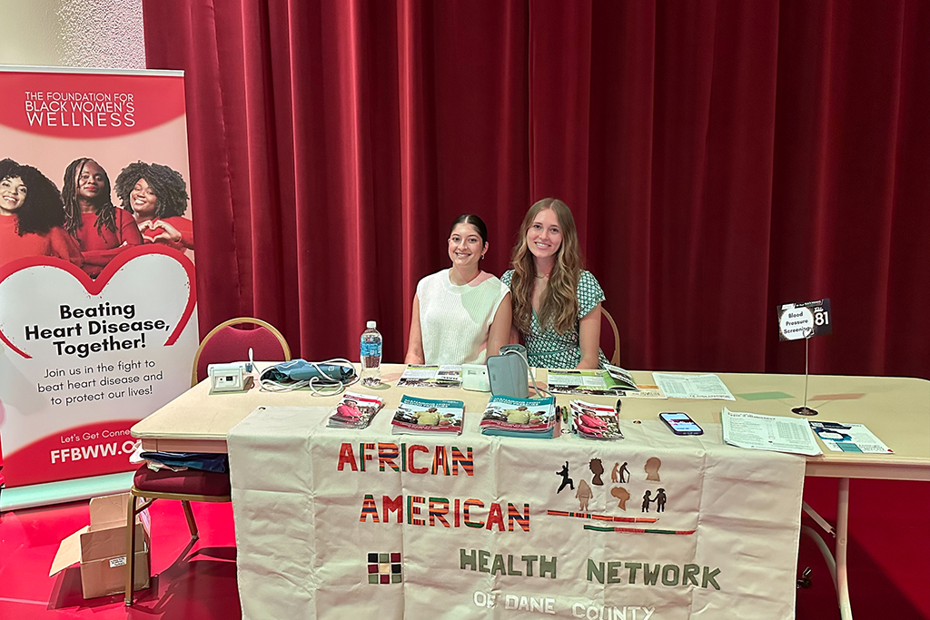 Jasmine Kuhnhenn and Makenzie Hansen sit at a table in front of a red background. The table has pamphlets, a blood pressure monitor, a glucose monitor, and a cloth that reads African American Health Network.