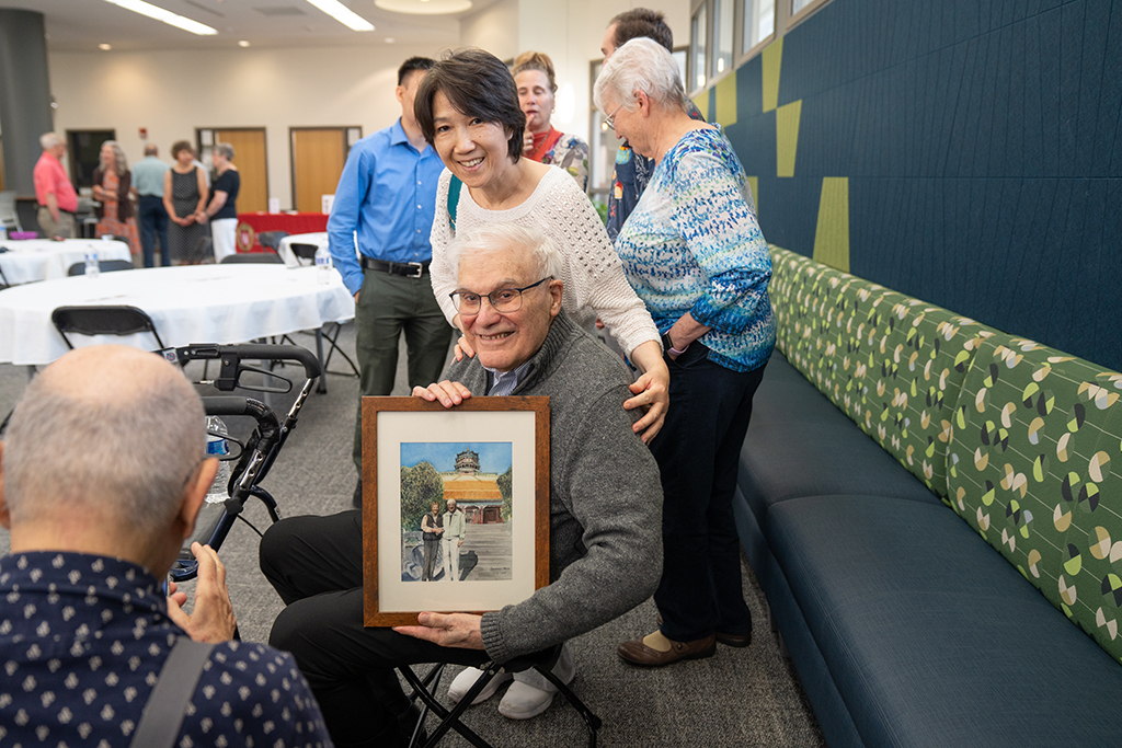 George Zografi poses with a drawing of two individuals in front of a building, with a woman standing behind him with her hand on his shoulder. A photographer is in front of George, snapping a picture.