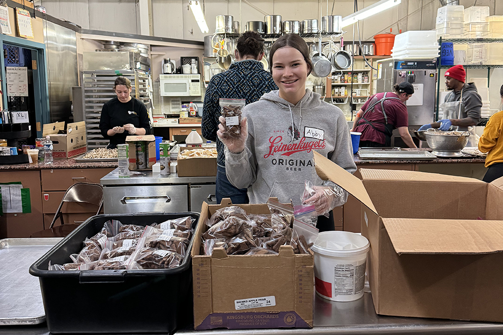 Abby Opsal holds up a bag of goods in an indoor setting surrounded by boxes of foods.