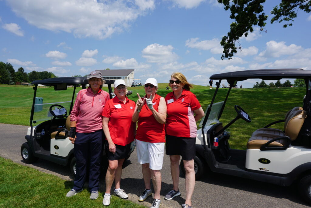 Four female alumni wearing red stand and smile in front of golf carts