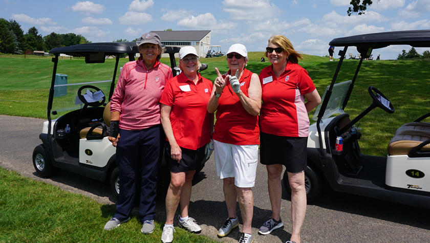 Four female alumni wearing red stand and smile in front of golf carts