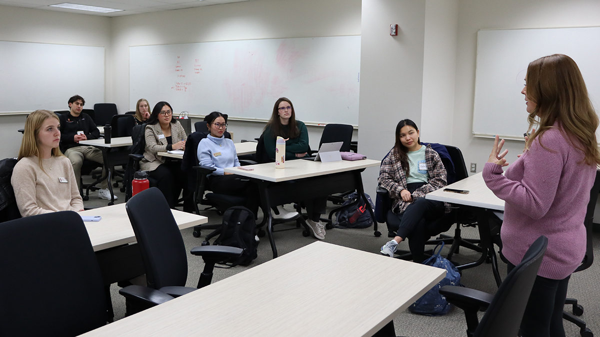 PharmD students seated at long tables listen to a speaker who is standing in front of them, facing away from the camera.