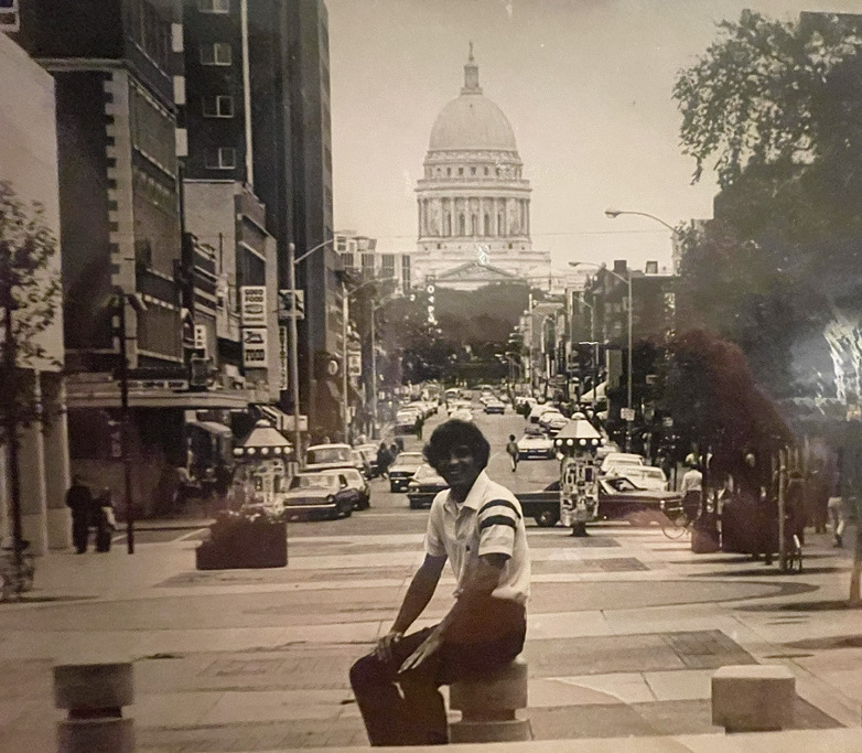 A back and white photo of a young man sitting on a concrete pillar with the Wisconsin State Capital in the background