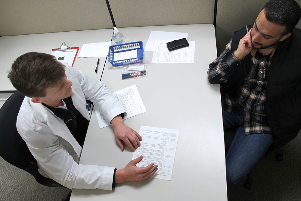A student in a white coat shares a handout with a standardized patient across a table.