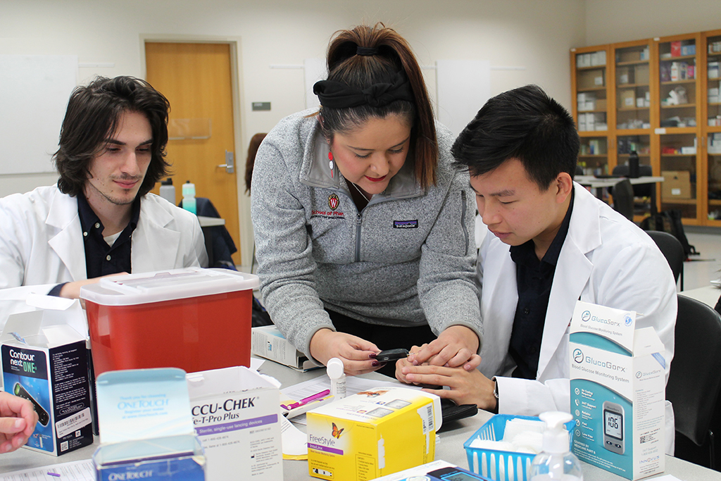Marina Maes helps two PharmD students practice using a glucose monitor.