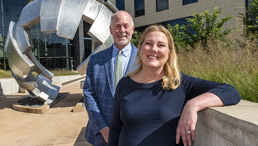 Lee Vermeulen and Jill Kolesar lean against a wall in front of a metal sculpture