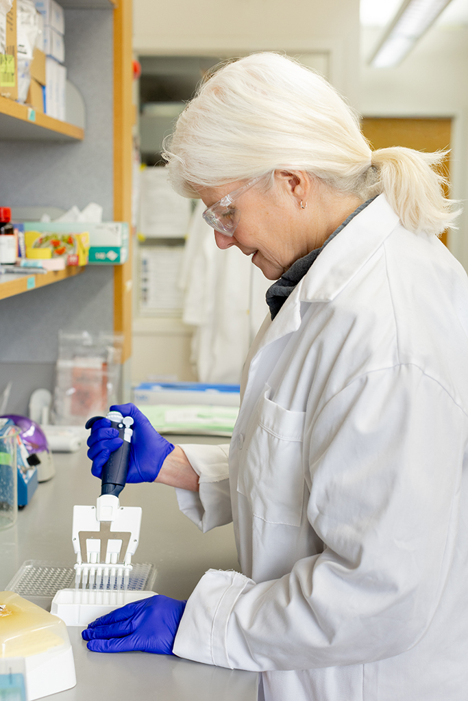 Sue McCrone holding a pipette while wearing gloves and a white coat