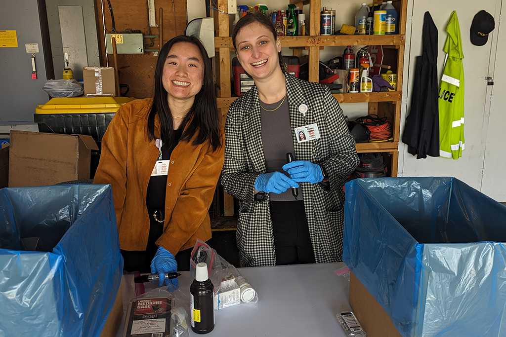 Two female PharmD students pose for a photo behind a table that's holding cardboard boxes lined with blue bags, a locking medicine case, and other assorted medication bottles.