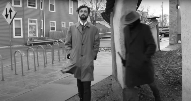 A black and white video still of a man walking with a man wearing a dark hat and coat approaches him.