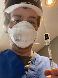 Nick Batinich in PPE, holding a vial of vaccine.