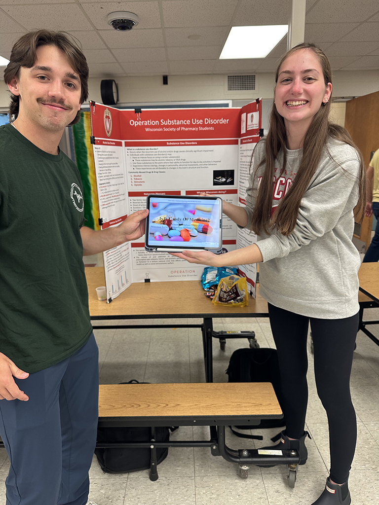 Lukas Kelsey and Jessica Haase hold a tablet that has a photo of pills on it in front of a poster about Operation Substance Use Disorder.