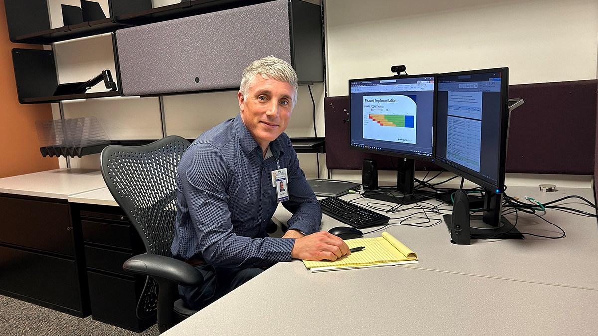 Tom Dilworth smiles for a photo while working at his desk with papers and a computer