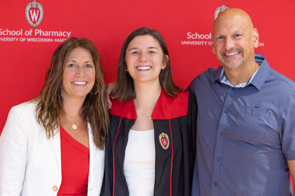 A PharmTox graduate poses with her family in front of the red School of Pharmacy backdrop.