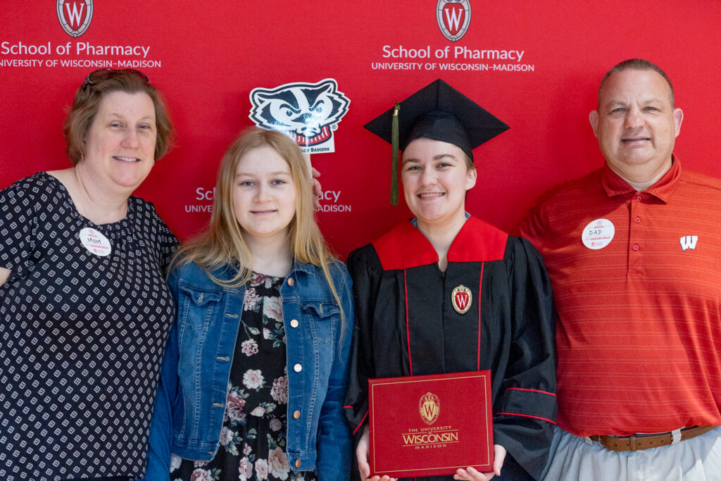 A PharmTox graduate and her family pose in front of the red School of Pharmacy backdrop.