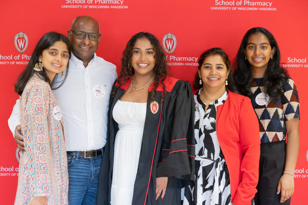A PharmTox graduate and her family pose in front of the red School of Pharmacy backdrop.