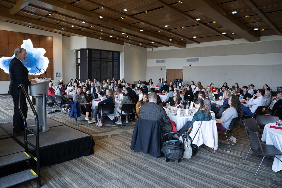 Dave Mott speaks at a microphone in front of tables of student and donor attendees.