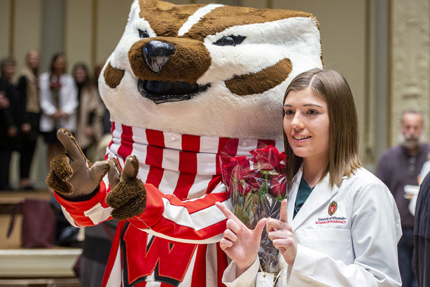 Student in white coat forming a 'W' with their hands with Bucky Badger and a bouquet of roses in their arm